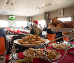 Platters laden with pastries, fruit mince tarts, bagels and croissants on a table in front of a very large dining table with many people seated around