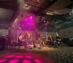 A group of musicians sitting in a semi-circle with various instruments underneath a bright purple disco light