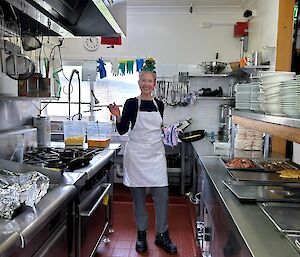 A smiling lady in a white apron stands in a commercial kitchen