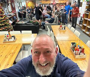 A smiling man takes a selfie with a group of people and a Christmas tree behind him
