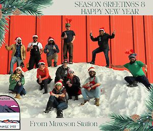 Small team on snow in front of red workshop building smiling at camera wearing Santa caps or reindeer horns, photo surrounded by Christmas message and Mawson crest