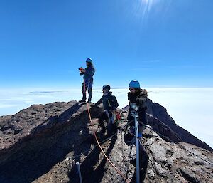 3 men harnessed for climbing on top of rocky peak with ice surrounding and into the distance, blue sky above