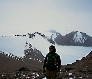 Man looks to camera from top of mountain with other mountain ranges and peaks in distance