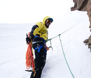 Man in climbing harness and ropes on snowy slope turns and smiles back at camera