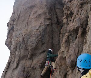 Two men dressed for climbing, look on as a man ascends rock face