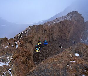 Looking down on hollow in rocks where four people are sheltering, around is foggy slopes