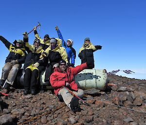 Group of seven people sitting on, or standing behind, 2 44 gallon drums, point away from camera in all different directions