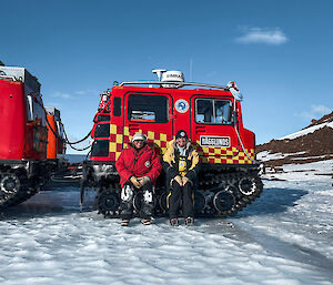 Two women in cold weather gear, sitting on side tracks of hagglunds vehicle, smiling to camera