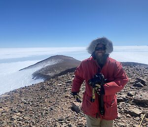 Man at top of long rocky slope, wearing goose down with hood up, smiles to camera