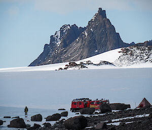 In the foreground, two Hägglunds parked with person walking across ice, in the distance large peak rising high into the sky