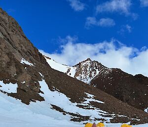 Camp site with yellow and red tents in the snow at the base of a step rocky mountain