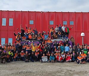 Large group of expeditioners in Christmas attire gathered together for a group photo in front of the Casey red shed