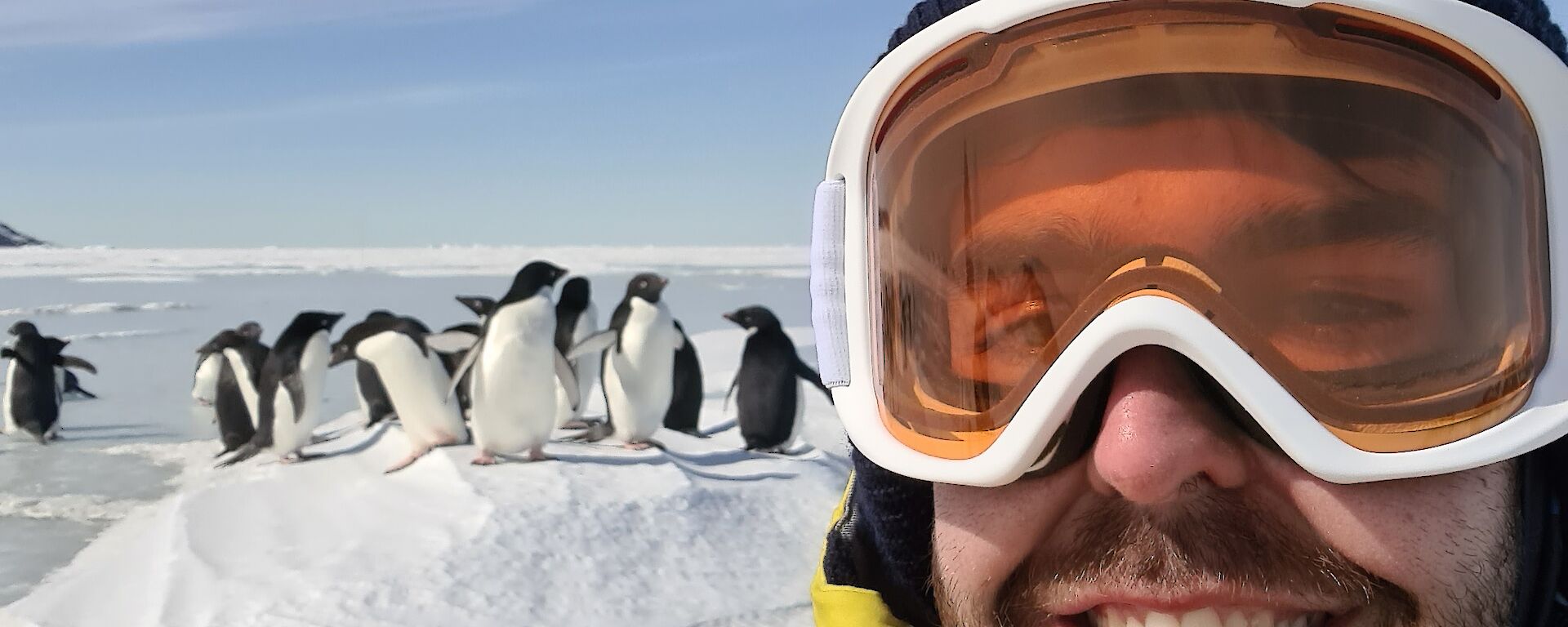 A smiling man in snow goggles and a beanie standing in front of a group of penguins in the snow