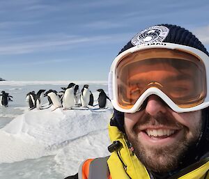 A smiling man in snow goggles and a beanie standing in front of a group of penguins in the snow