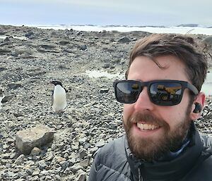 A smiling man in sunglasses stands in front of a sole penguin that has wandered over to take a look
