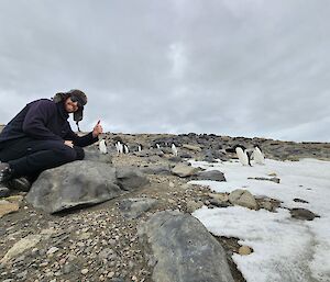 A smiling man with a thumbs up kneels on a rock with a group of penguins in the background
