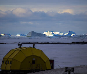A half-cylinder shaped, yellow building in the foreground with the sea-ice in the background. The horizon in the distance shows icebergs illuminated by shafts of sunlight coming through the clouds in the distance.