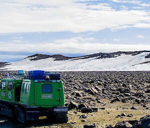 A green box-shaped vehicle with a box-shaped trailer both mounted on tracks parked amongst boulders in the foreground with snow-covered hills in the distance
