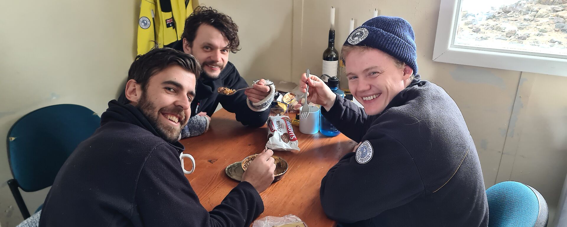 Three smiling men, each holding a spoon, sitting around a table sharing a pie from a tin