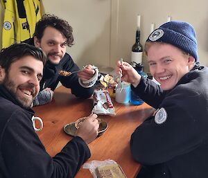 Three smiling men, each holding a spoon, sitting around a table sharing a pie from a tin