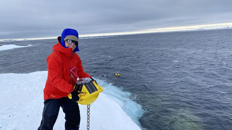 A woman in a red jacket holds a scientific instrument over sea ice with the ocean behind her