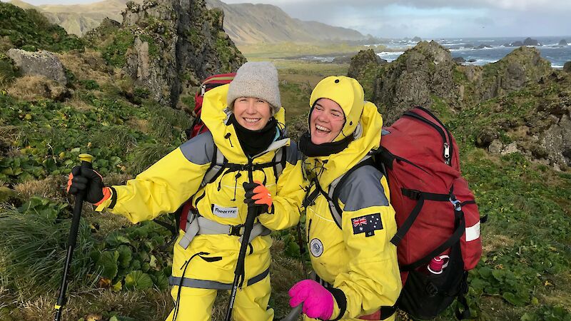 Two women in yellow survival clothing smile, with Macquarie Island behind them
