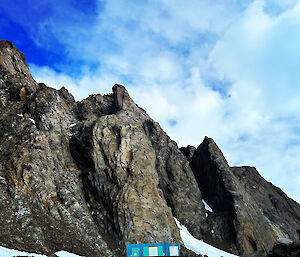 Small rectangular blue hut with wooden balcony sits at the base of rocky mountain range