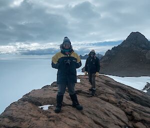 Two men stand on ridge of rock with drops away sharply either side, in the background another rocky peak and the ice plateau