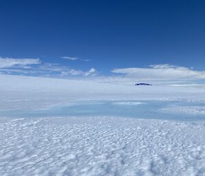 Expanse of ice with blue sky above, and in dip of landscape a lake of melted water has formed