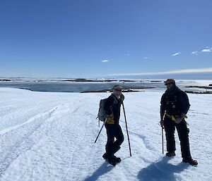 Two men in foreground holding ice axes and walking stick, one ice slope with water and islands in the background, clear blue skies above