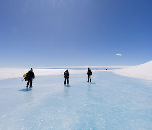 Three people walking away from camera across a slick blue ice lake with sun and blue sky above