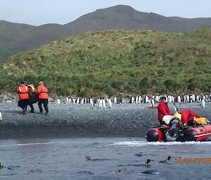 Unloading a red rubber boat and carrying gear up a grey beach as penguins stand by watching