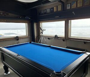 A blue pool table in the foreground with three windows in the background with spectacular views of the sea-ice