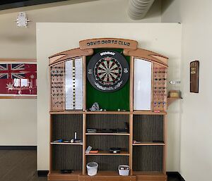 A wall with a dart board and surrounded by a dart board cabinet with shelves, storage for darts and whiteboards for scoring.