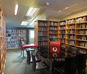 A room filled with bookshelves and books with a table in the middle surrounded by chairs. The table has a small red cardboard postbox on it.