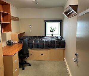 A small bedroom with a window, bed, desk and chair and shelving units.