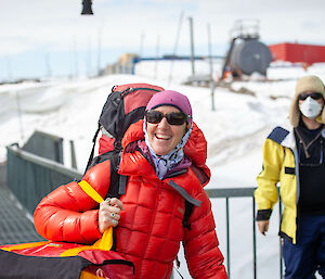 A lady in a red jacket and carrying a backpack and a red survival bag sports a huge grin.
