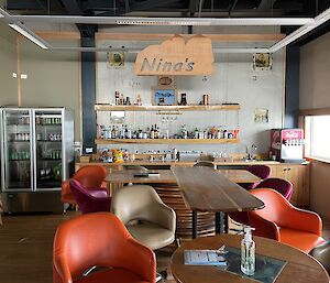 A bar with a long table and bar stools with a drinks fridge, bottle of drinks on shelves, and a soft drink machine in the background