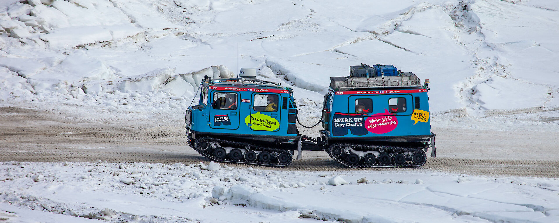 A blue vehicle with a trailer, both on tank tracks driving up a dirt road surrounded by ice and snow. People can be seen inside the vehicles.