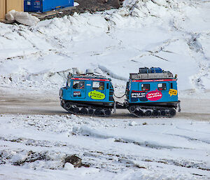 A blue vehicle with a trailer, both on tank tracks driving up a dirt road surrounded by ice and snow. People can be seen inside the vehicles.