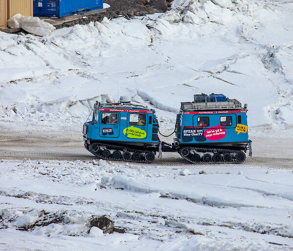 A blue vehicle with a trailer, both on tank tracks driving up a dirt road surrounded by ice and snow. People can be seen inside the vehicles.