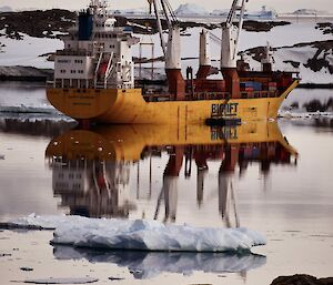 Large yellow cargo vessel reflected in calm water against a backdrop of rocks and snow