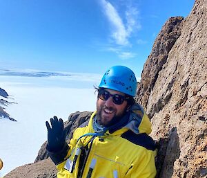 Close up of man in climbing helmet and yellow shell jacket at top of mountain waves to camera