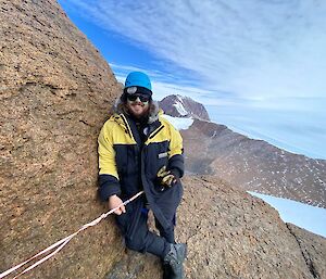 Close up of man in climbing helmet and AAD doona jacket, harnessed with ropes, at top of mountain smiles to camera