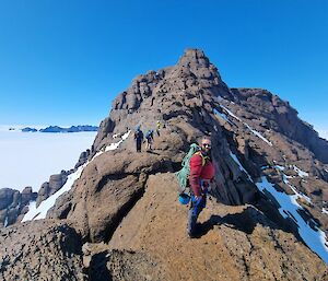 Man standing on ridge line of mountain, carrying ropes and climbing helmet, with main peak in background, smiles to camera