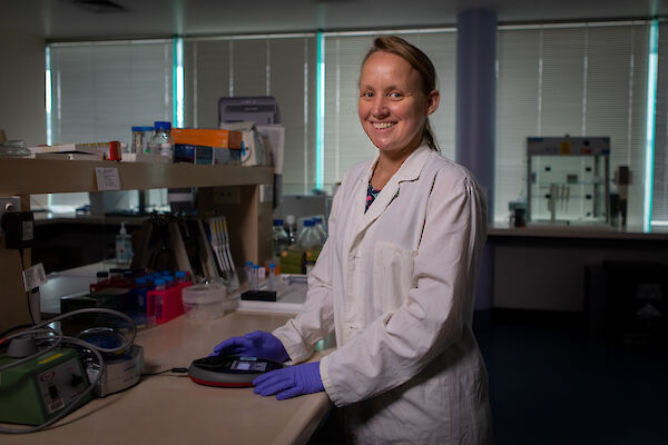 A woman in a white lab coat standing at a laboratory bench, smiling at the camera.