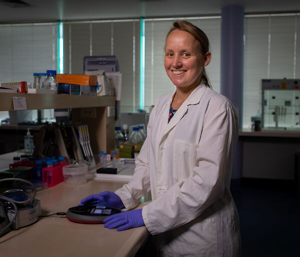 A woman in a white lab coat standing at a laboratory bench, smiling at the camera.