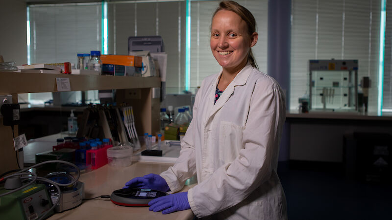 A woman in a white lab coat standing at a laboratory bench, smiling at the camera.