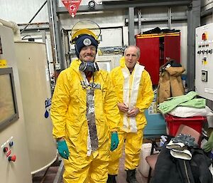 Two smiling men wearing yellow protective suits and black workboots.