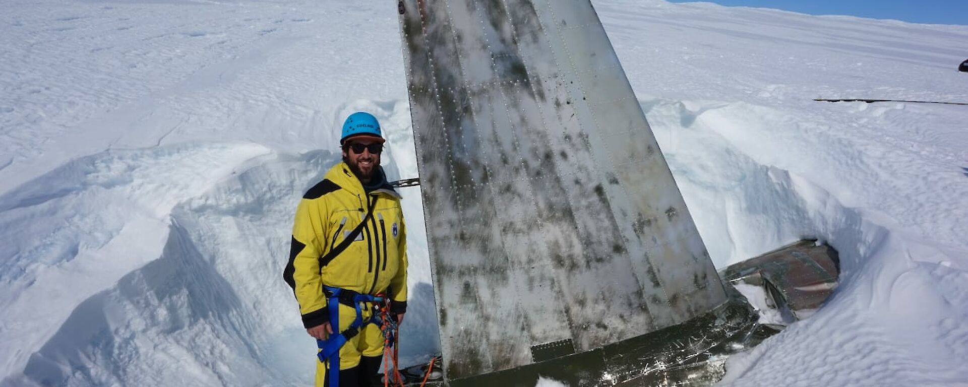 Man in harness and safety helmet stands beside aircraft tail which is rising up out of the ice